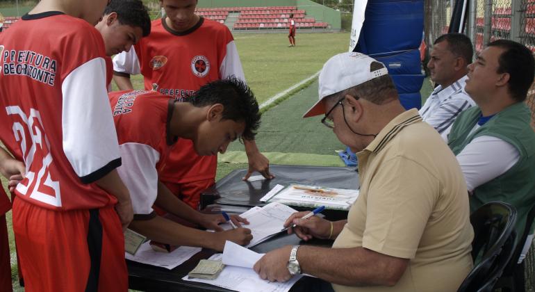 Mesário, acompanhado dois auxiliares, observa jogadores assinarem um livro registrando presenças.