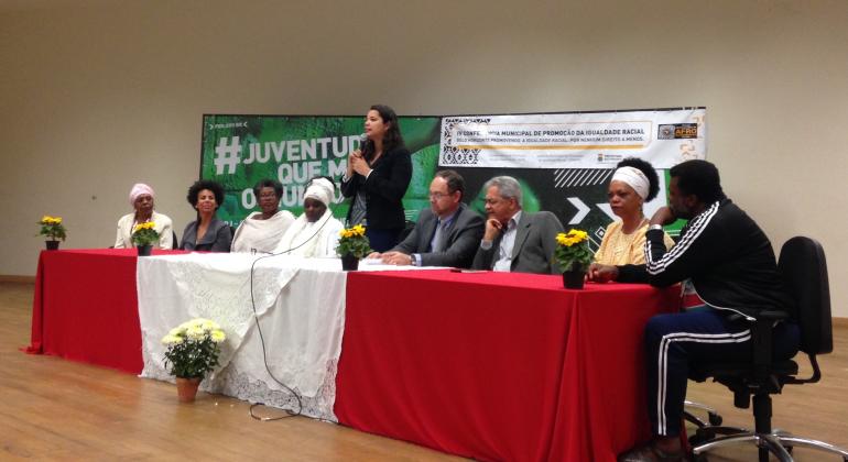 Nove pessoas compondo a mesa de abertura da Conferência de Igualdade Racial; com destaque para a secretária municipal de Políticas Sociais Maíra Colares de pé, ao centro.