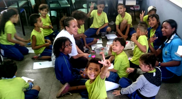 Cerca de treze crianças realizam dinâmica com orientadora, sentados no chão. 