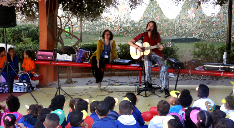 Semana Nacional da Educação Infantil foi celebrada na região Oeste com apresentações artísticas, passeata, teatro e folclore. Foto: Fabiana Rosa/PBH Mulher faz apresentação musical de violão para mais de vinte crianças, ao fundo, outra mulher observa.