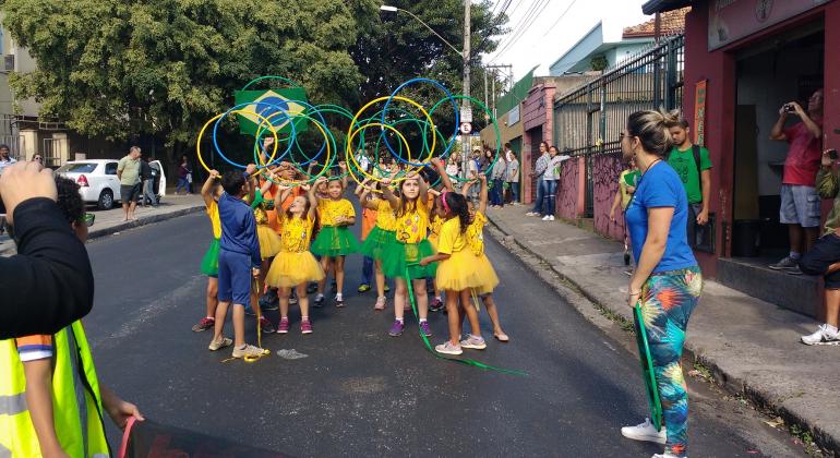 Mais de dez meninas de camiseta amarela e saias verdes ou amarelas e um menino de bermuda e casaco azul fazem movimentos com bambolês verdes, amarelos ou azuis em passeata na rua. Junto ao grupo, ainda havia a bandeira do Brasil.