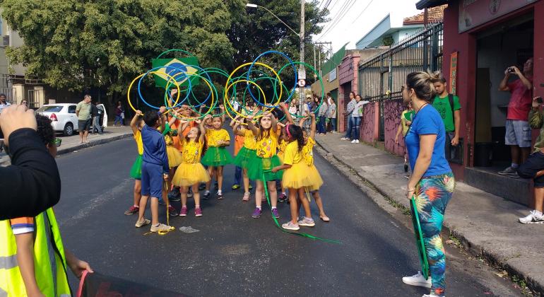 Foto: Iara Souza Mais de dez meninas de camiseta amarela e saias verdes ou amarelas e um menino de bermuda e casaco azul fazem movimentos com bambolês verdes, amarelos ou azuis em passeata na rua. Junto ao grupo, ainda havia a bandeira do Brasil.