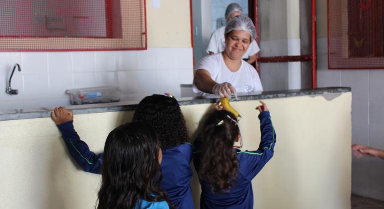BH em Pauta: Aplicativo auxilia supervisores de alimentação Três meninas na fila para a merenda enquanto uma funcionária entrega uma banana para uma delas