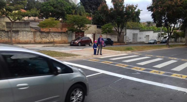 Mulher, de mão dada com criança, atravessa faixa de pedestre em rua de bairro. 
