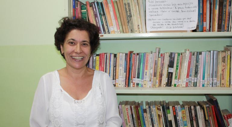 Centro de Saúde Vera Cruz, na região Leste, cria biblioteca para aproximar as pessoas da literatura e da arte. Foto: Divulgação PBH MUlher sorri à frente de estante com livros.