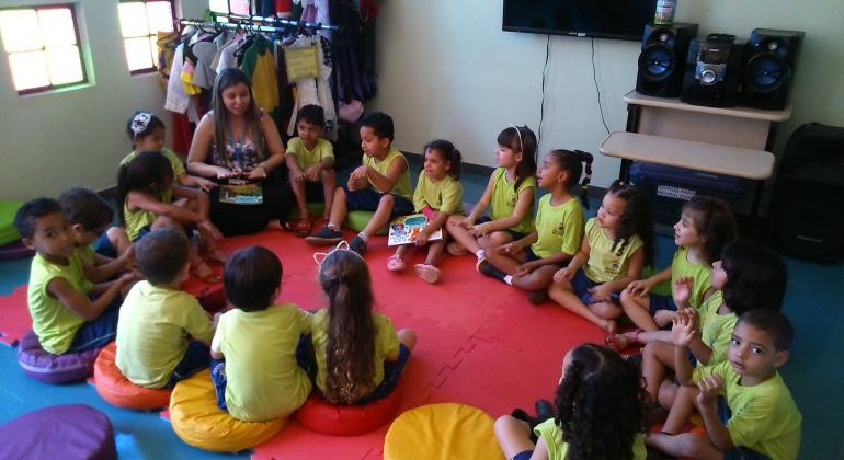 Trabalho desenvolvido em Unidade Municipal de Educação Infantil desperta as crianças para o universo literário. Foto: Mara Damasceno/PBH Treze crianças sentadas escutam história de professora.