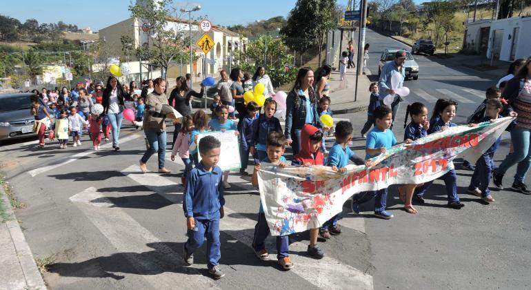 Mais de vinte crianças com menos de sete anos, acompanhadas de professores, nas ruas da Pampulha com cartaz sobre a Semana de Educação Infantil