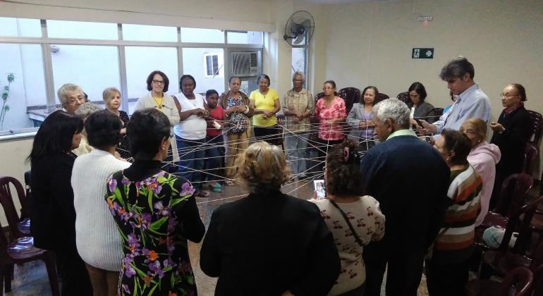 Foto: Divulgação PBH Cerca de catorze idosos em círculo seguram barbantes que formam uma rede, conectando todos as todos.
