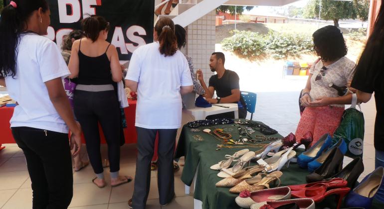 Três mulheres escolhem itens como sapatos e bijuteiras em uma banca com dois atendentes