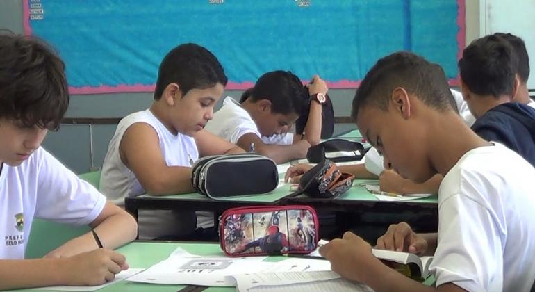 Mais de seis alunos, sentados dois a dois em mesas, fazendo provas ou testes.