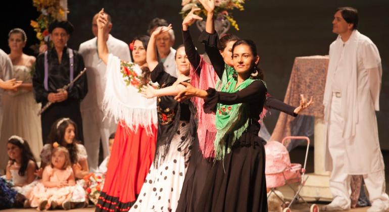 Obra literária "Nosso Lar", de autoria Chico Xavier, foi adaptada para o teatro e será exibida no Raul Belém Machado. Foto: Divulgação Quatro mulheres com saias longas, blusas de mangas compridas e chales dançam à frente; ao fundo, homem todo de branco à direita e à esquerda, homens, mulheres e crianças assistem.