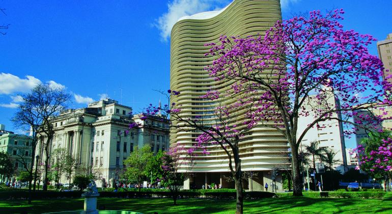 Praça da Liberdade com ipê rosa, Edifício Niemeyer e Centro Cultural Banco do Brasil. À frente, fonte com laguinho circular. 