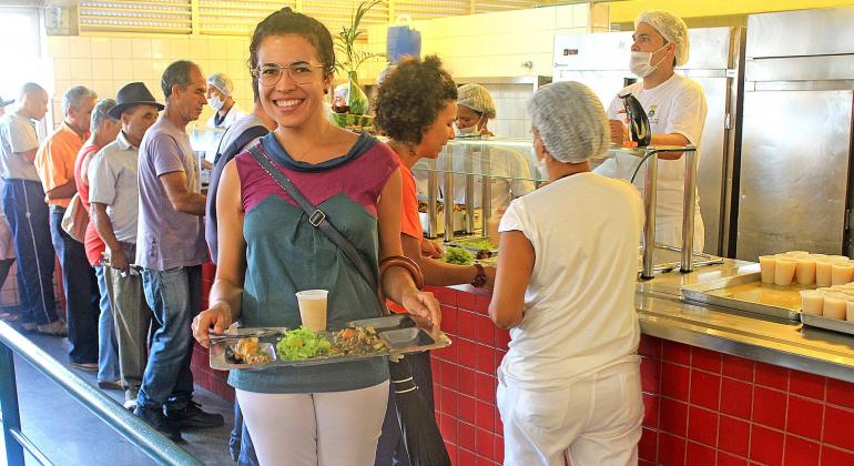 Cidadão sorri com bandeja de almoço em Restaurante Popular; ao fundo, fila para almoço com cidadãos e atendentes.