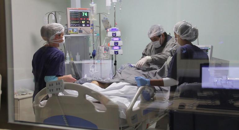 Sala de hospital médico e dois profissionais de saúde realizando procedimentos em paciente na cama com equipamentos médicos.