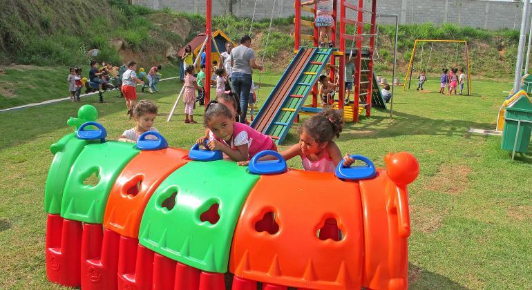 Cerca de quinze crianças, acompanhadas por educadores infantis, em pátio de Umei; uma brincam em túnel colorido, outras em escorregador e outras em cadeiras de balanço.