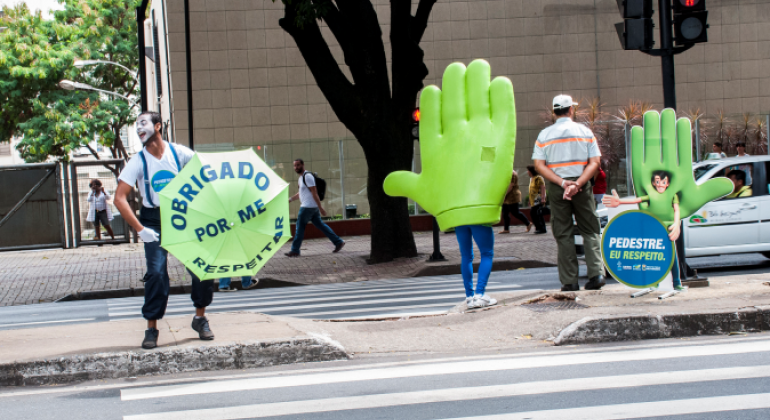 Campanha de trânsito é realizada em faixa de pedestre. Dois personagens: um mímico segurando um guarda chuvas escrito "Obrigado por me respeitar" e uma mão verde, que representa o "pare" do semáforo. Além de um guarda municipal de costas.