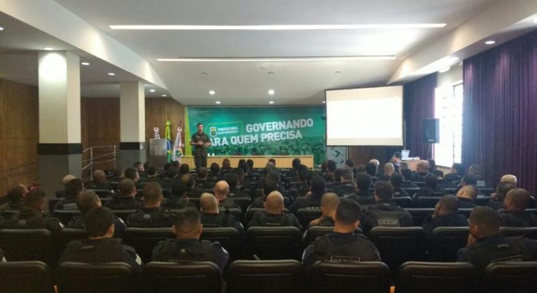 Agentes da Guarda Municipal participam de curso do Exército Grupo de cerca de 80 guardas estão sentados em um auditório, de costas para a foto. No palco, de frente, está um membro do exército brasileiro. Um banner escrito "Governando para quem precisa" está ao fundo.
