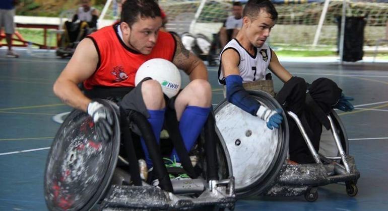 Foto: Divulgação Dois jogadores de rugby em cadeiras de rodas em ação em quadra de esportes.