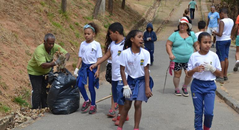 Quatro alunos de escola municipal caminham no Parque Vila Pinho durante o dia; ao lado, funcionário recolhe lixo; atrás, cidadãos também fazem caminhada. 