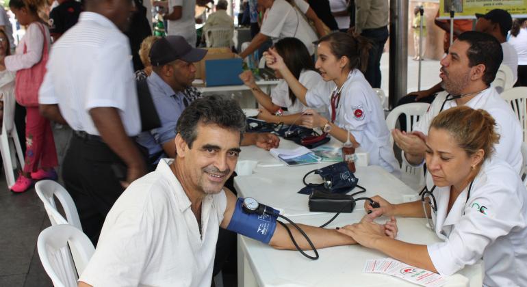 Cidadão sorrindo, em destaque, recebe atendimento de pressão. Ao fundo, outros atendimentos