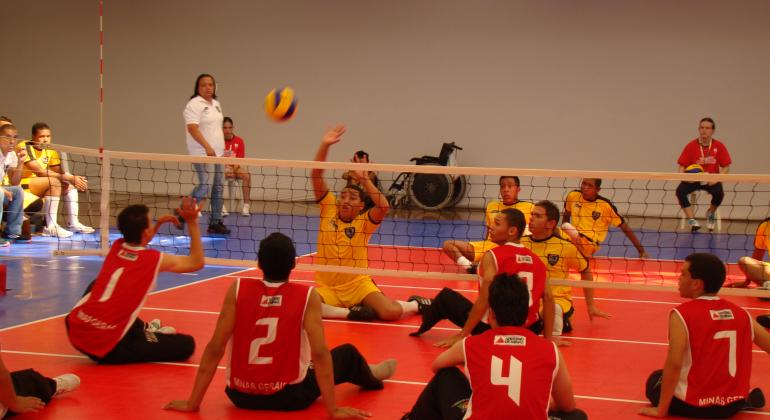 Jogo de vôlei com jogadores sentados: Na frente, time com uniforme vermelho e atrás, time com uniforme amarelo. A bola está no ar. 