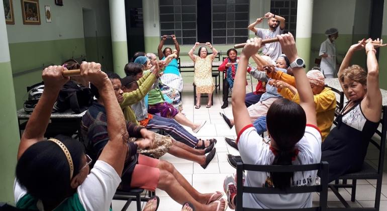 Cerca de dez idosos se exercitam, sentados e com as mãos para cima, segurando minibastões, orientados por instrutor que está de pé. 