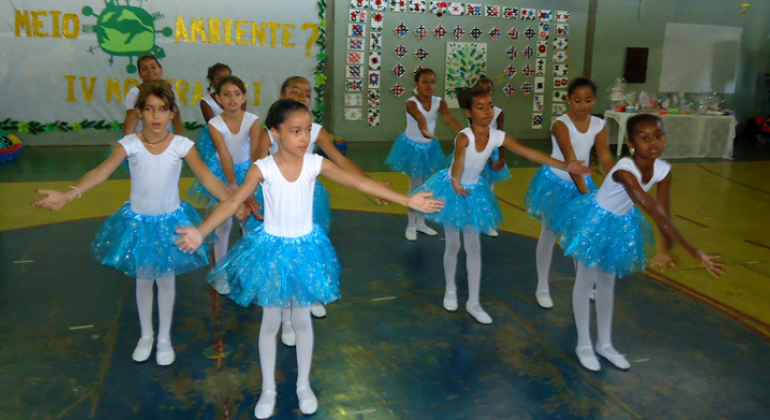 Dez alunas com roupas de balé apresentam coreografia na quadra da escola.