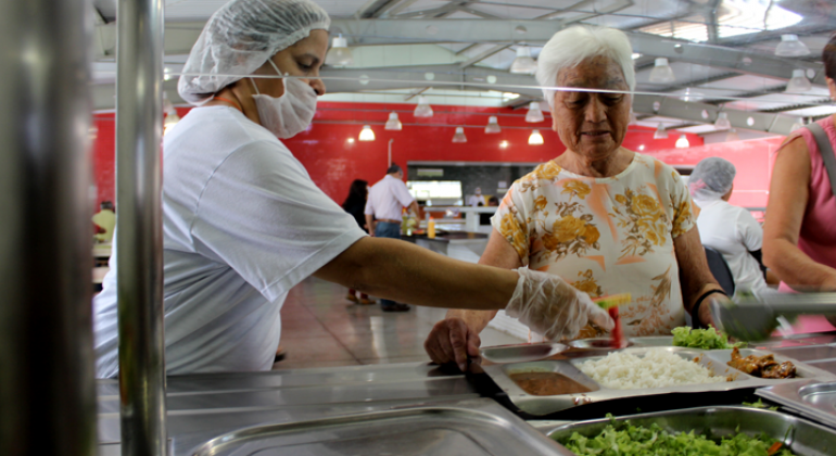 Refeições oferecidas nos quatro restaurantes populares de Belo Horizonte são escolhidas e preparadas sob rígidos padrões nutricionais e de sabor. Foto: Divulgação/PBH Funcionária do restaurante popular servindo refeição à uma senhora.