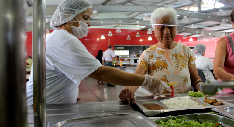 Funcionária do restaurante popular servindo refeição à uma senhora.