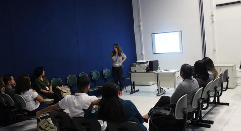 BH em Pauta: Oficina melhora o atendimento à comunidade LGBT Instrutora, de pé, fala para oito servidores da prefeitura de Belo Horizonte, sentados em meia lua. O ambiente é uma sala fechada. Há cinco cadeiras vazias.