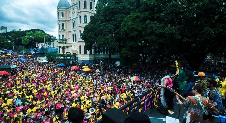 Prefeitura lança edital de patrocínio para o Carnaval - Foto: Danilo Candombe/Acervo Belotur Bloco de carnaval Beiço do Wando, no Carnaval de 2017. Foliões usam roupas rosa e amarelo. Há um prédio ao fundo e uma grande árvore.