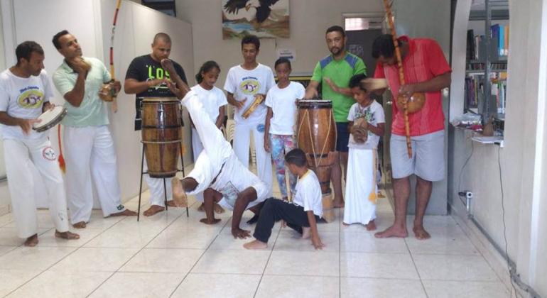 Roda de Capoeira com instrumentos musicais e onze pessoas; duas delas jogam capoeira ao centro.
