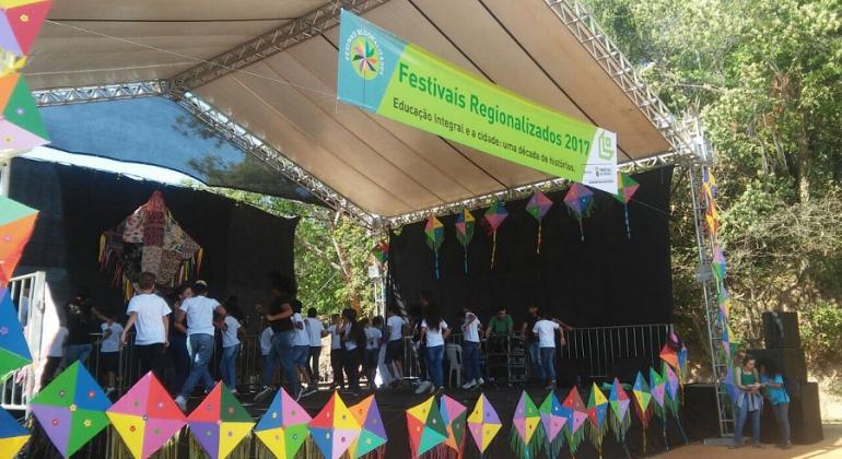 Em outubro, quatro as regionais Barreiro, Leste, Norte e Oeste realizam Festivais Regionalizados e comemoram 10 anos do Programa Escola Integrada; encontros vão até dezembro. Foto: Welington Guimarães/PBH Mais de dez pessoas se apresentam em palco coberto ao ar livre; no alto do palco, uma faixa com os dizeres "Festival Regionalizado 2017"