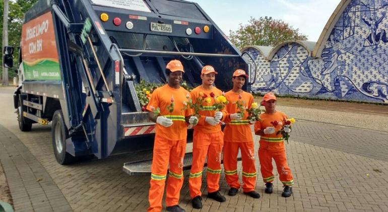 Garis e agentes da SLU distribuirão rosas a pedestres e motoristas dias 24 e 25 de outubro na Pampulha e na praça da Liberdade. Foto: Divulgação PBH Quatro garis seguram rosas em frente ao caminhão de recolhimentop de lixo, tendo ao lado a Igrejinha da Pampulha.