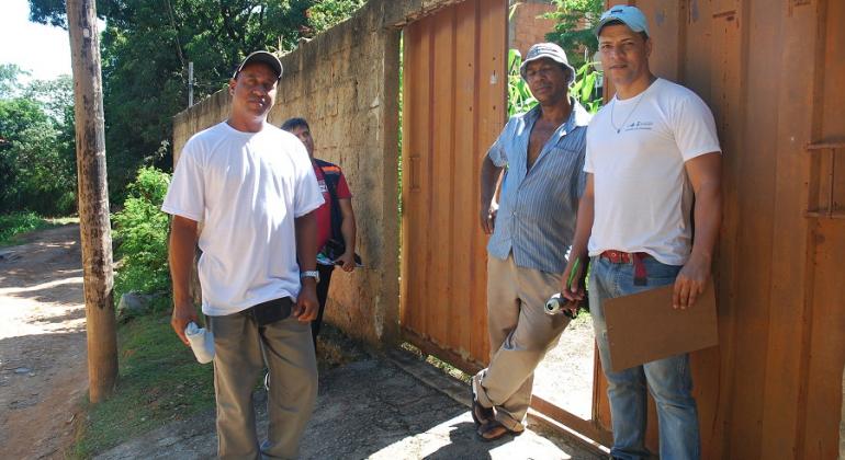 Dois agentes comunitários de saúde no portão de uma residência, junto ao proprietário, durante o dia. 