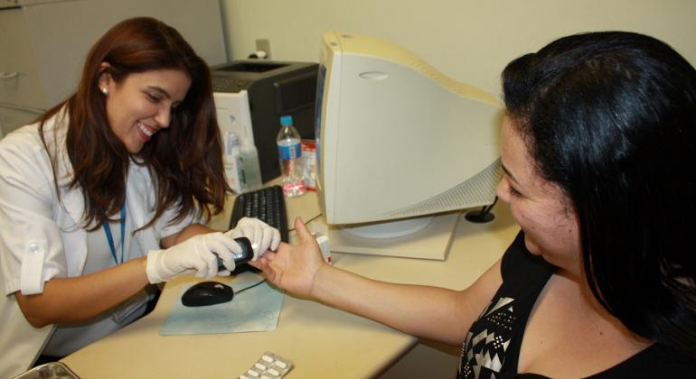 Todos os 152 centros de saúde de BH contam com atuação de farmacêutico de referência, o que amplia os desfechos clínicos favoráveis. Foto: Divulgação PBH Cidadã mede glicemia em centro de saúde; uma cartela de remédio pode ser vista em uma mesa.