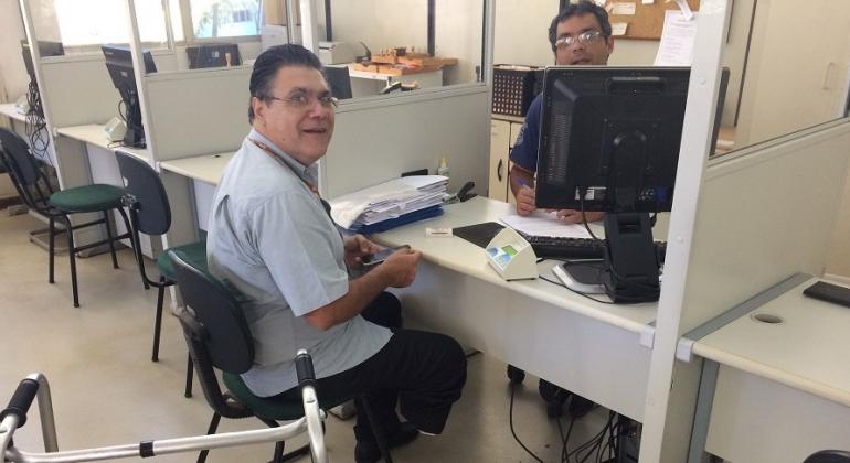 Saiba como solicitar serviços e informações sobre trânsito, ônibus e gratuidade de transporte em Belo Horizonte. Foto: Divulgação PBH Homem solicita serviços da BHTrans em atendimento presencial; ele está sentado em frente a uma mesa, do outro lado, um homem o atende com computador.
