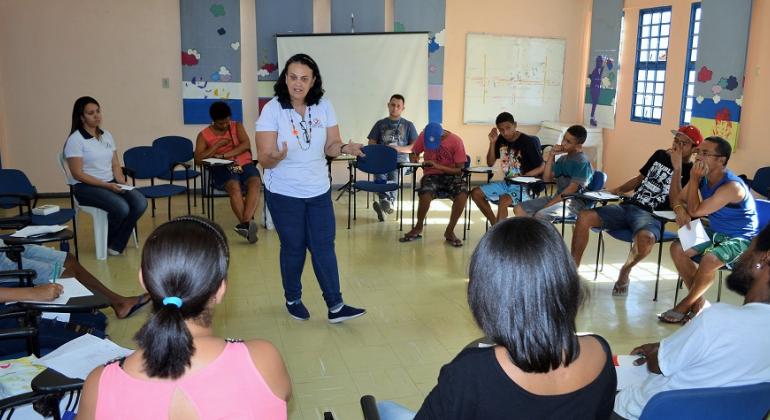 Em área de vulnerabilidade social na região Noroeste, Prefeitura desenvolve cursos e oficinas voltados a jovens com idade entre 14 e 19 anos. Foto: Sidney Procópio/PBH Cerca de treze jovens, sentados e em círculo, assistem a aula de instrutora que está de pé, em uma sala, durante o dia.