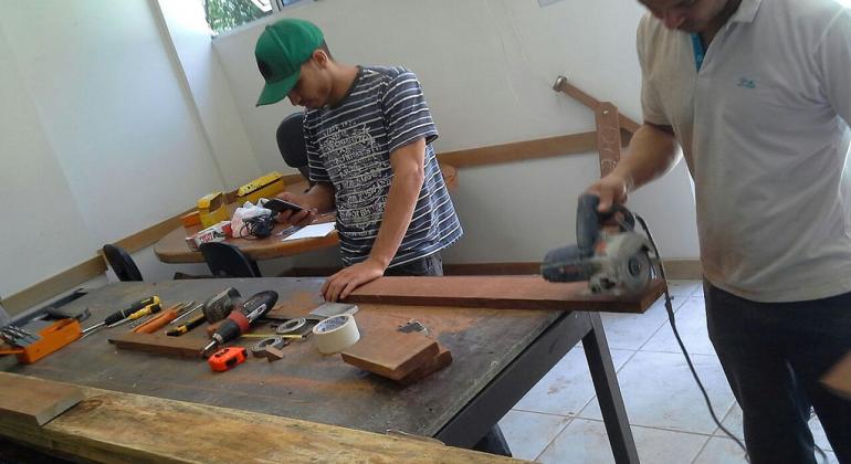 Projeto com campeonato de carrinho de rolimã mobiliza alunos de 20 escolas municipais da Regional Norte de Belo Horizonte. Foto: Geraldo Félix/PBH Um homem e um jovem de boné constróem carrinho de rolimã em oficina.
