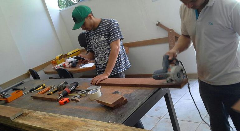 Um homem e um jovem de boné constróem carrinho de rolimã em oficina. 