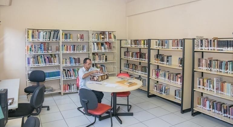 Homem sentado em mesa em biblioteca com várias estantes de livros. 