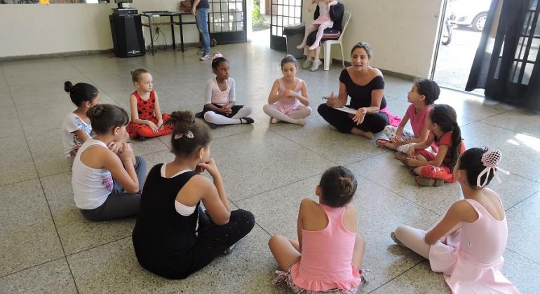 Centro Cultural Pampulha oferece espaço para manifestações artísticas e culturais em área de vulnerabilidade social. Foto: Divulgação PBH Nove garotas fazem aula de balé, sentadas em círculo, orientadas por professora; ao fundo da sala, uma garota está sentada no colo de responsável.