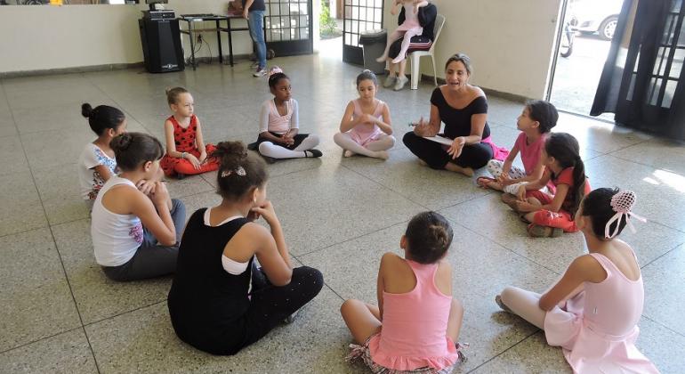 Nove garotas fazem aula de balé, sentadas em círculo, orientadas por professora; ao fundo da sala, uma garota está sentada no colo de responsável. 