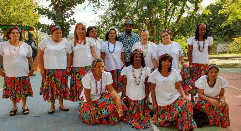 Onze umlheres participantes do grupo de canto Cigarras da Vitória, com blusa branca e saia florida; ao centro, homem com camisa azul e branca e chapéu.