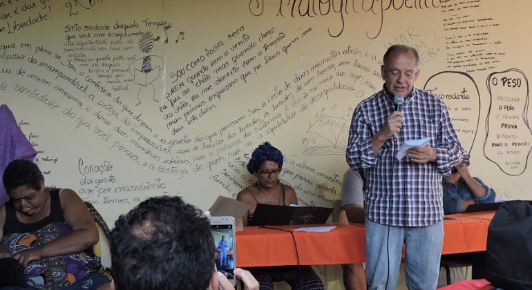 Homem declama poesia à frente de um mural com vários escritos. 