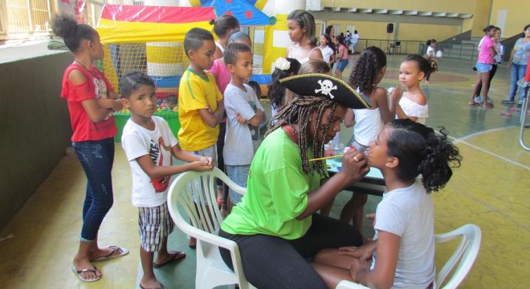 Mulher com chapéu de pirata maquia uma criança, outras nove estão ao redor dela. 
