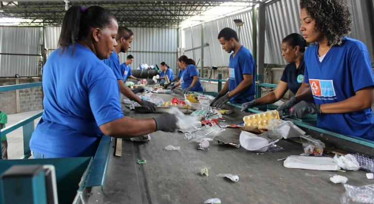 Oito pessoas fazem coleta seletiva de lixo utilizando uma esteira elétrica. 