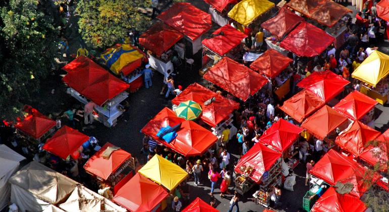 Vista aérea da Feira de Artes e Artesanato da Avenida Afonso pena, com cobertura das barraquinhas vermelhas em destaque e pessoas circulando entre elas. 