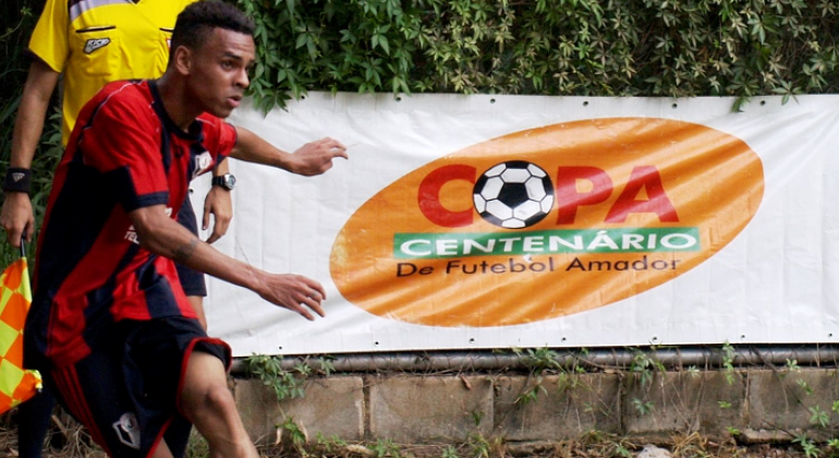 Jogador de futebol amador durante partida, com o banner da "Copa Centenário" ao fundo.