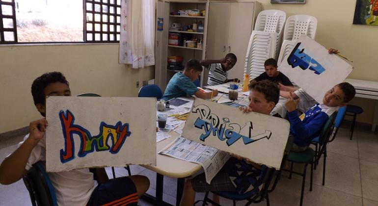 Cinco alunos, sentados em mesa, fazem oficina de grafite; três deles mostram seus desenhos coloridos. 